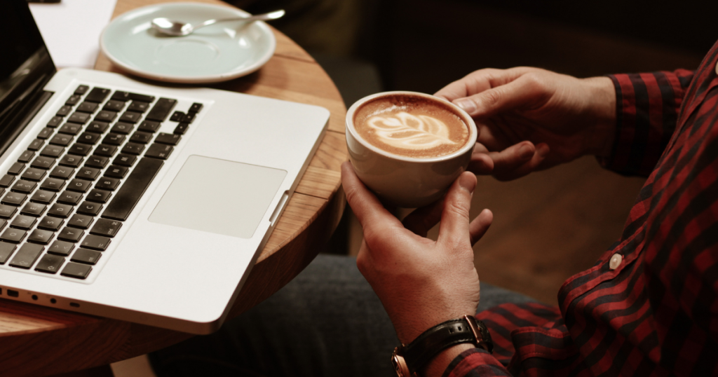 Working with Laptop from Coffee Shop by Jovana Stojanovic _ Getty Images - Genuine Origin Coffee A person holding a cup of coffee in their hands at a coffee shop table with an open laptop on the table.