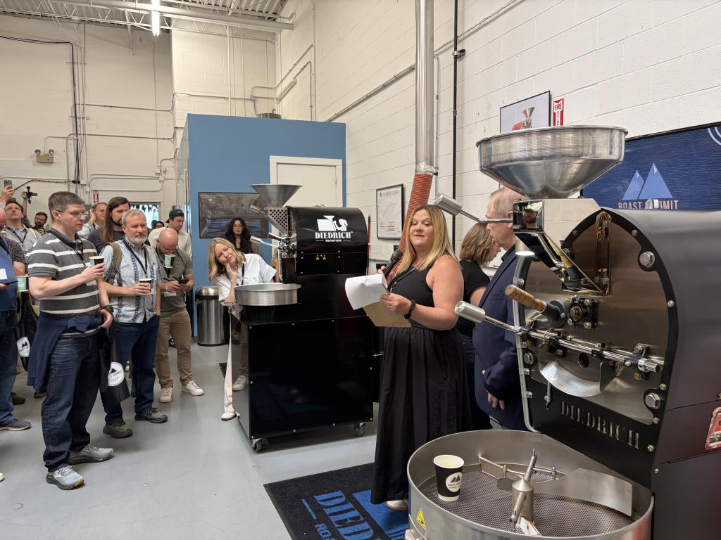 A group of people watches a coffee roasting demo beside a large Diedrich roasting machine in an industrial workspace. 
