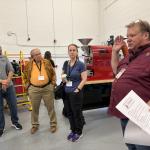 Group of five adults with name badges listening to a presenter beside a red coffee roaster in a workshop setting.