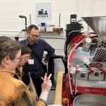 People gathered around a red Diedrich coffee roaster in a workshop, observing and discussing the machine.