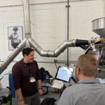 Group of people with lanyards gathered around a large industrial coffee roaster during a workshop, with metal ducts, a hopper, and control panel nearby.