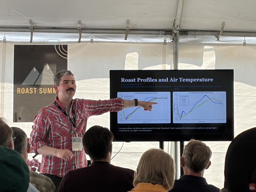 Presenter in a red plaid shirt points to a large screen showing 'Roast Profiles and Air Temperature' with roast graphs, in a tented venue.
