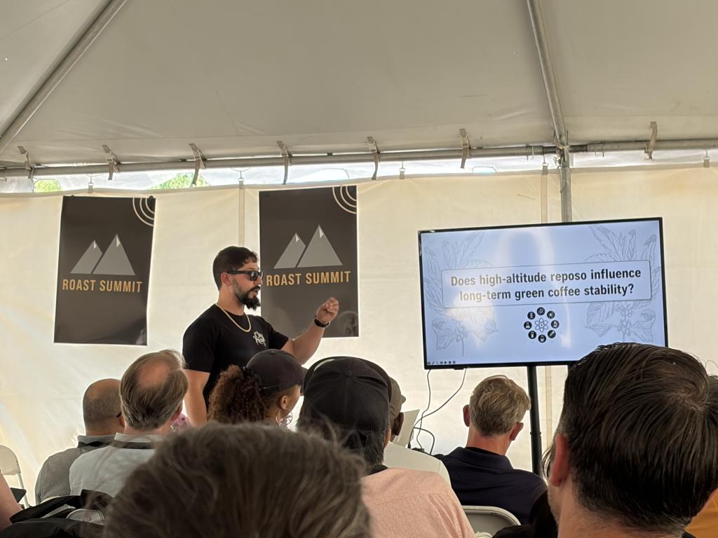 Speaker presenting at a Roast Summit in a tent, addressing a seated audience with a large screen displaying a coffee-themed slide statement about high-altitude effects on green coffee stability.
