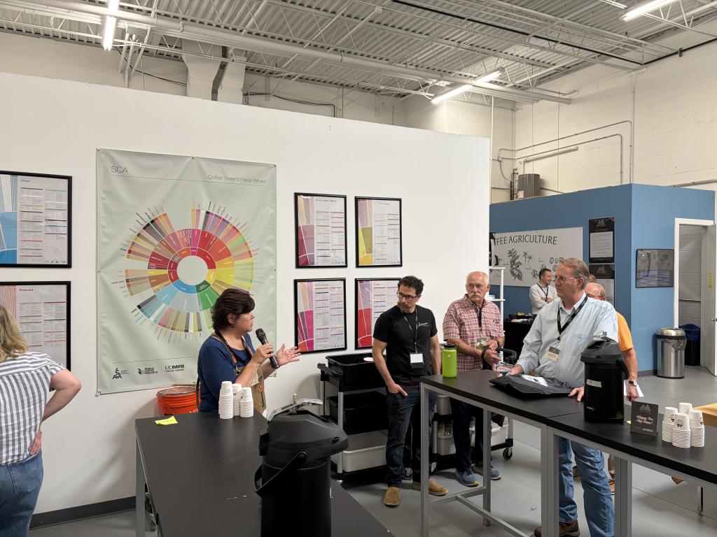 Renee Espinoze from Firedancer speaks into a microphone at a coffee-tasting booth with color-wheel posters on the wall behind her.