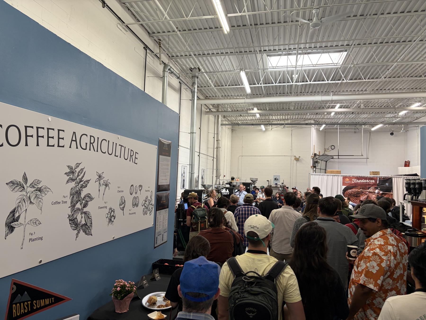 Crowd of people at a Roast Summit 2026, standing around a large poster titled 'COFFEE AGRICULTURE' on a blue wall.