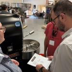 Two people examine a large black coffee roaster; a man with glasses holds a booklet while a woman in a striped shirt watches.