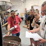 People gathered around a large coffee roasting machine, watching as roasted beans tumble inside the drum during a demonstration.