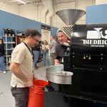 Man in a light shirt pours beans from a red bucket beside a Diedrich Roasters coffee roaster while others chat in the background.