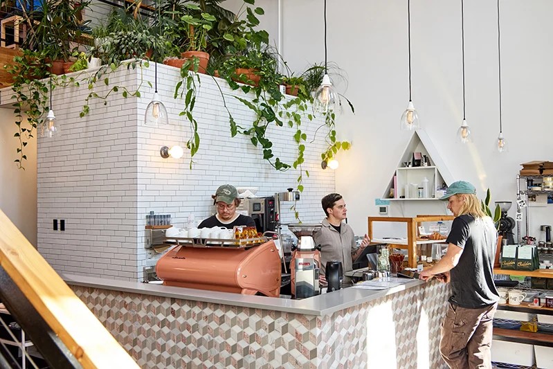 A barista in a clean and modern cafe talking to a customer behind a bronze espresso machine 