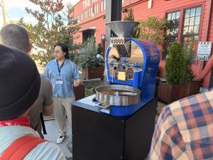 Speaker standing next to a blue Diedrich roaster as people listen to a presentation