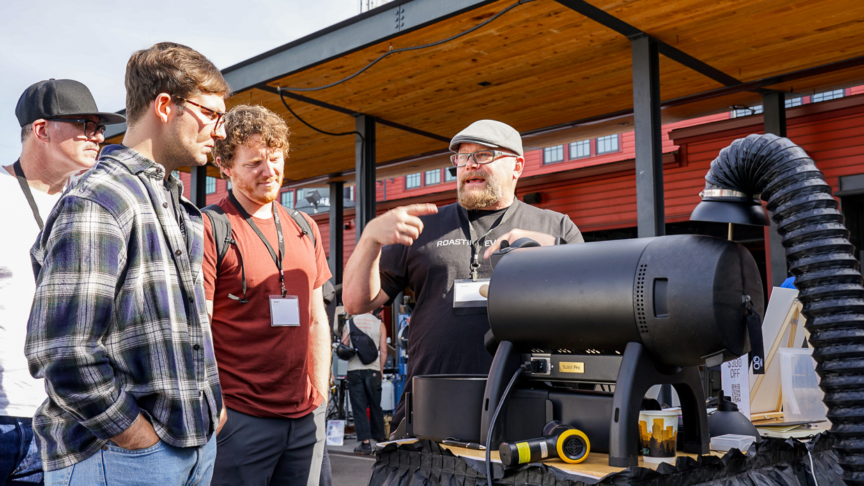 Three men listening to a roaster talk in front of a black coffee roaster outside