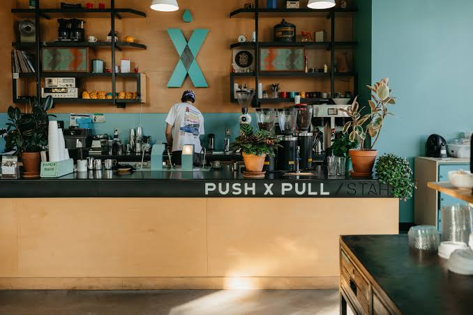 A nordic cafe with push x pull written on the bar and a barista with their back turned under a blue X logo